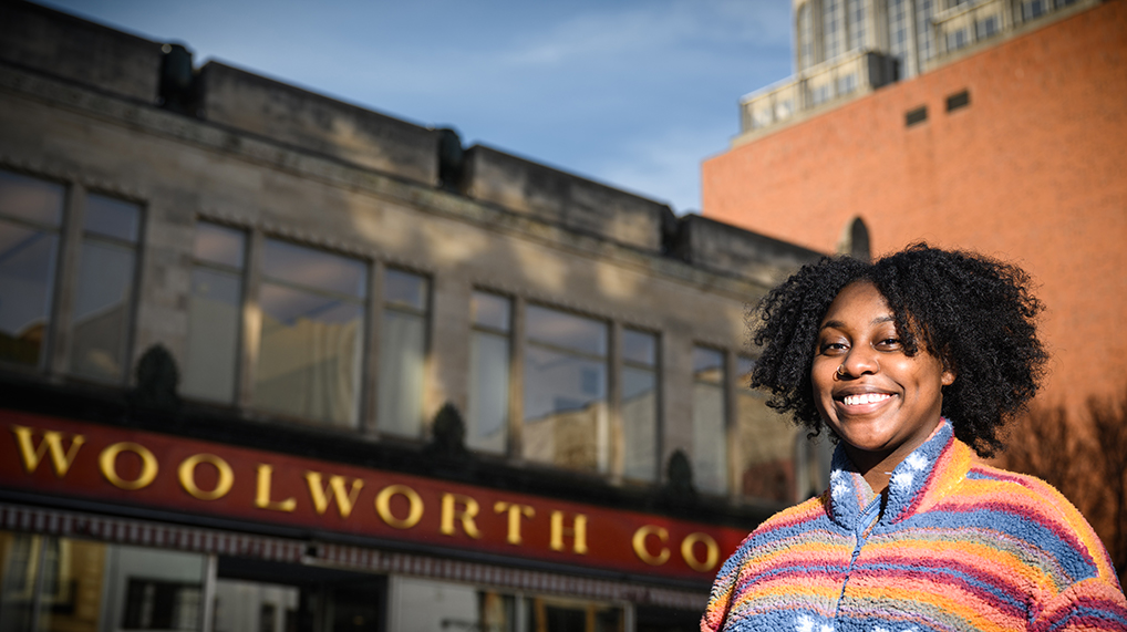 young Black woman stands in front of Woolworth Co building in Downtown Greensboro