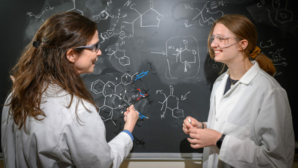 Professor Kimberly petersen stands at chalkboard with females student. Both wear labcoats. Chemistry equations are written on the board