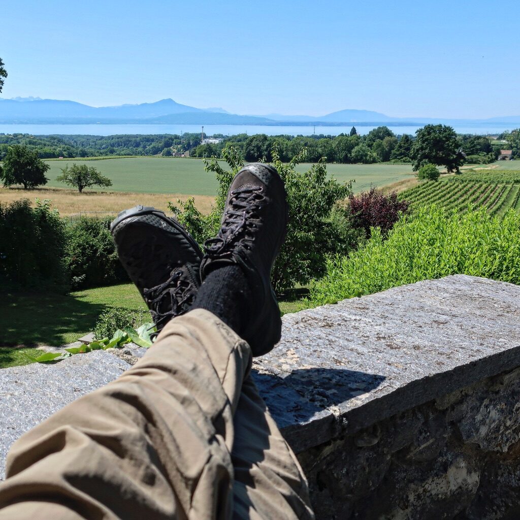 the view of a person's feet kicked up on a ledge overlooking green fields, a large body of water, and distant mountains and hills