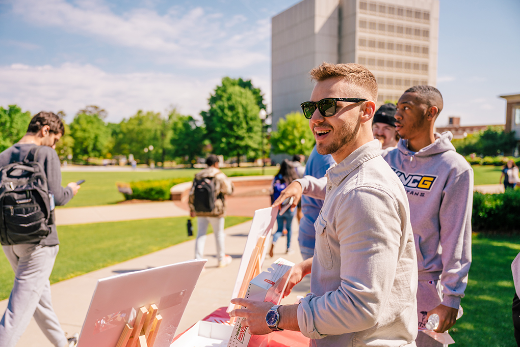 Ben Lambe, wearing sunglasses, hands out donuts to students walking past tables on campus. 