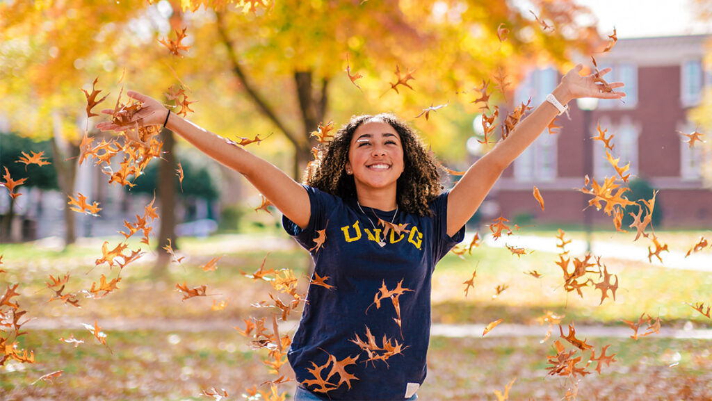 a student in a UNCG shirt frolics in golden fall foliage