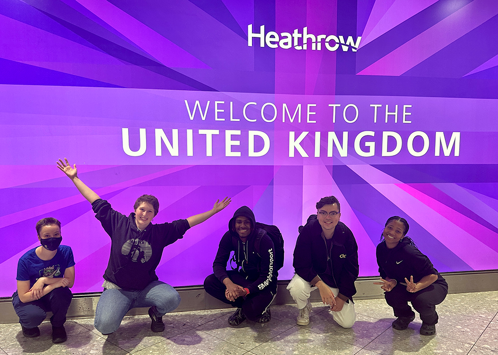 students crouched in front of Welcome to the United Kingdom sign at Heathrow airport