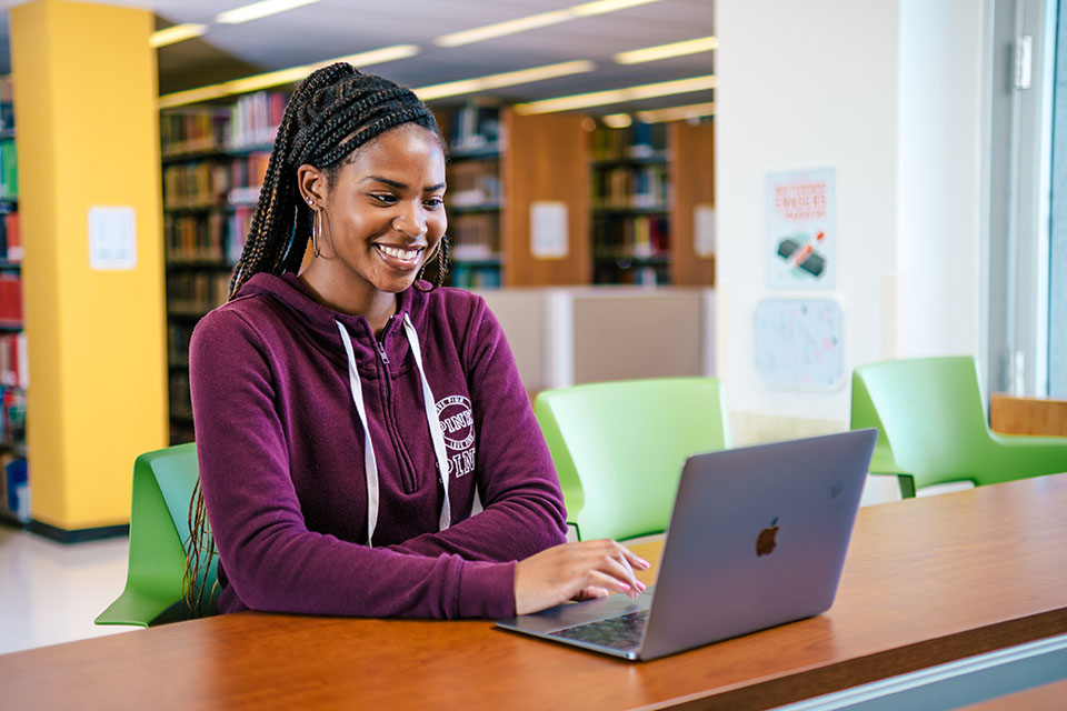 a student smiles while working on her laptop in a library setting.