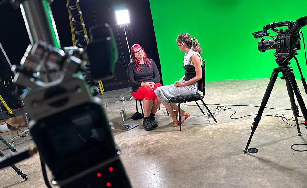 two women talk in front of green screen with camera equipment around