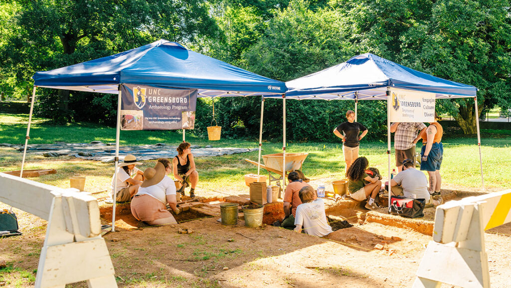 Students dig for artifacts in measured plots of an empty lot. Tents cover them with UNCG archaeology banners hanging and barricades mark off the site.