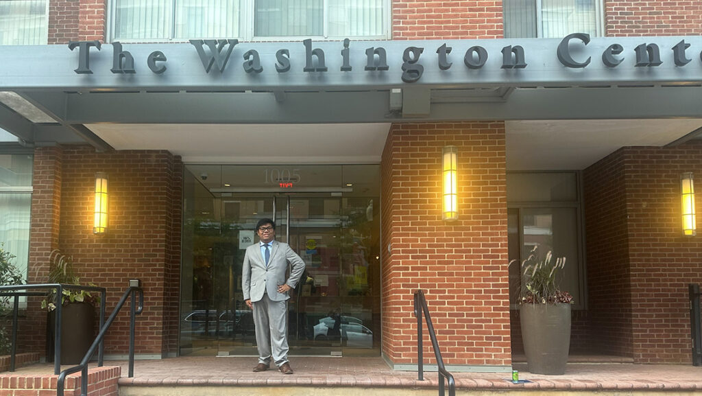 Student intern wearing suit stands in front of building with sign that says "The Washington Center"