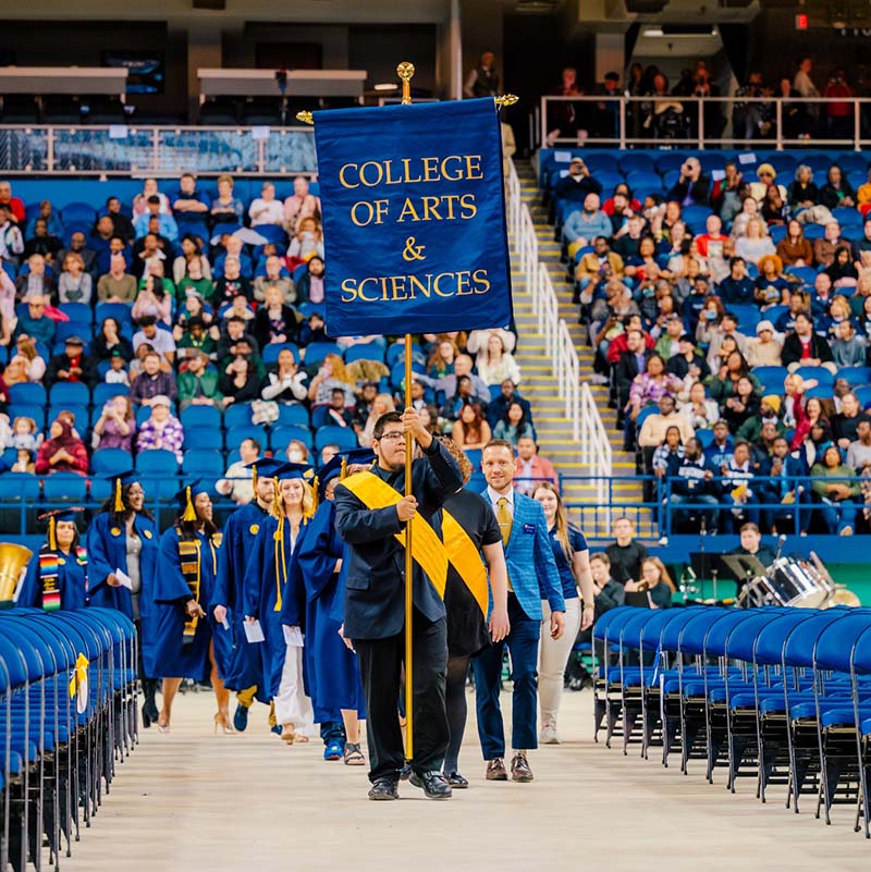 At graduation ceremony, student leads line of graduates while holding sign that says "College of Arts & Sciences"