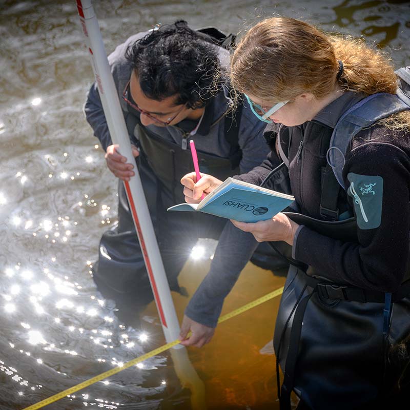 Geography Student and professor stand in river taking measurements with instruments