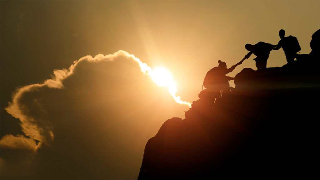 Group of people struggling to climb mountain with golden sun peeking behind clouds