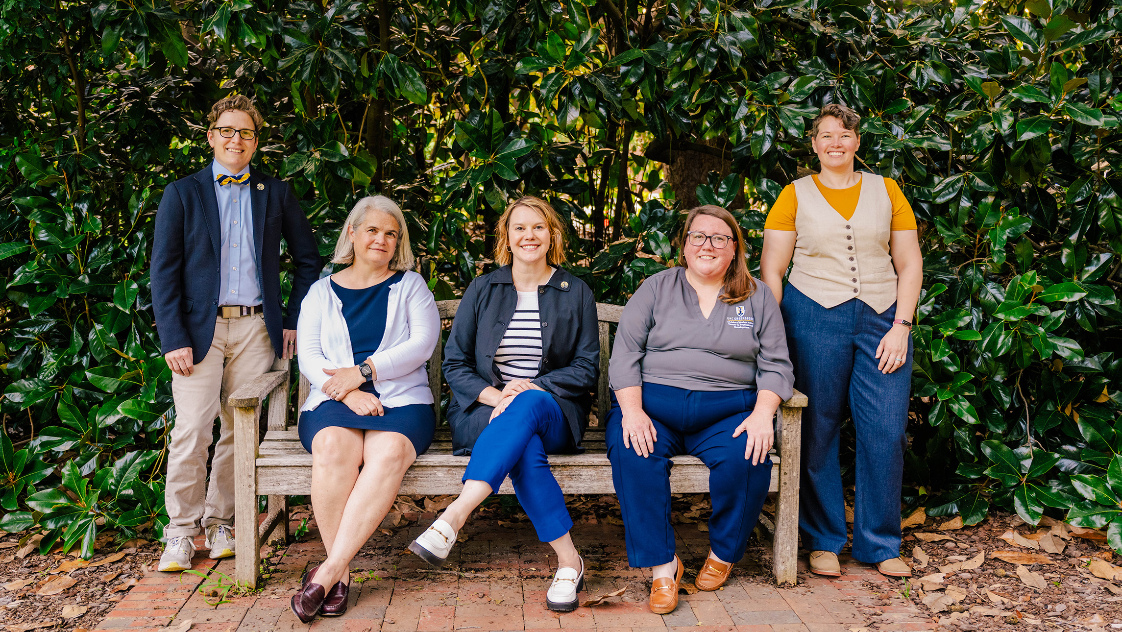 Humanities at Work team sits on and stands around a bench with greenery behind