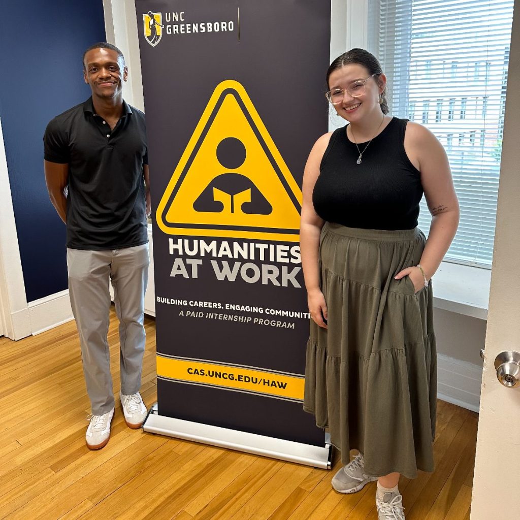 Two Humanities at Work team members stand with a pull-up banner with the Humanities at Work logo