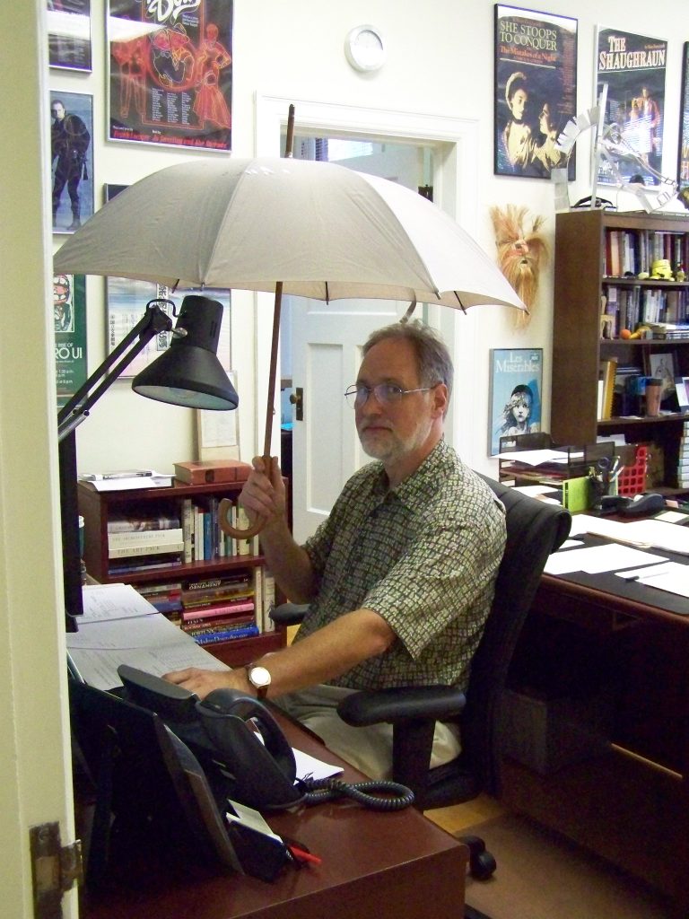Dr. Bob Hansen seated at a desk, holding an open umbrella over his head.