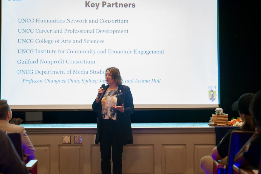 Dr. Heather Adams stands in front of a projected screen with a slide in the background that lists the Key Partners of Humanities at Work.