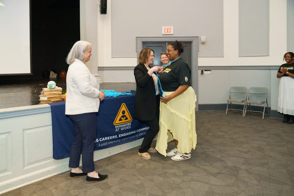 A Humanities at Work Intern receives a set of graduation cords from Dr. Heather Adams, while Dr. Maura Heyn looks on in the foreground.
