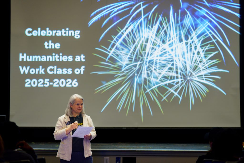 Dr. Maura Heyn stands in front of a projected screen with the words "Celebrating the Humanities at Work Class of 2025-26" with and an image of fireworks.