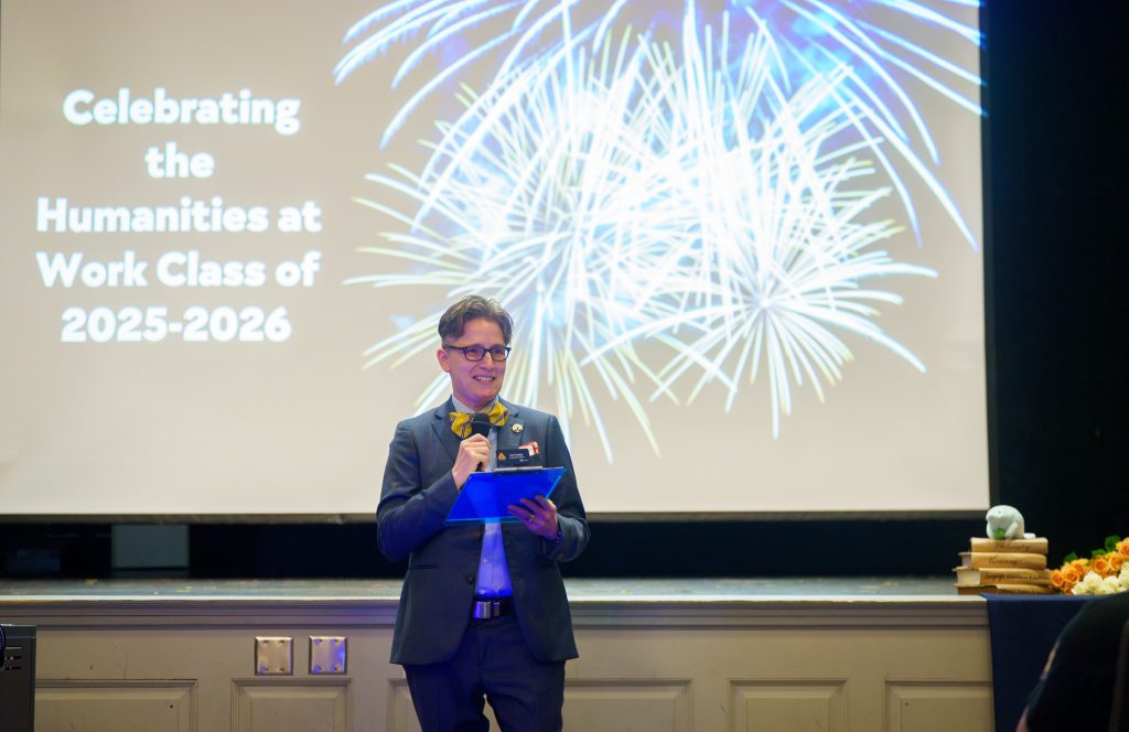 Dr. Jennifer Feather stands in front of a projected screen with the words "Celebrating the Humanities at Work Class of 2025-26" with and an image of fireworks.