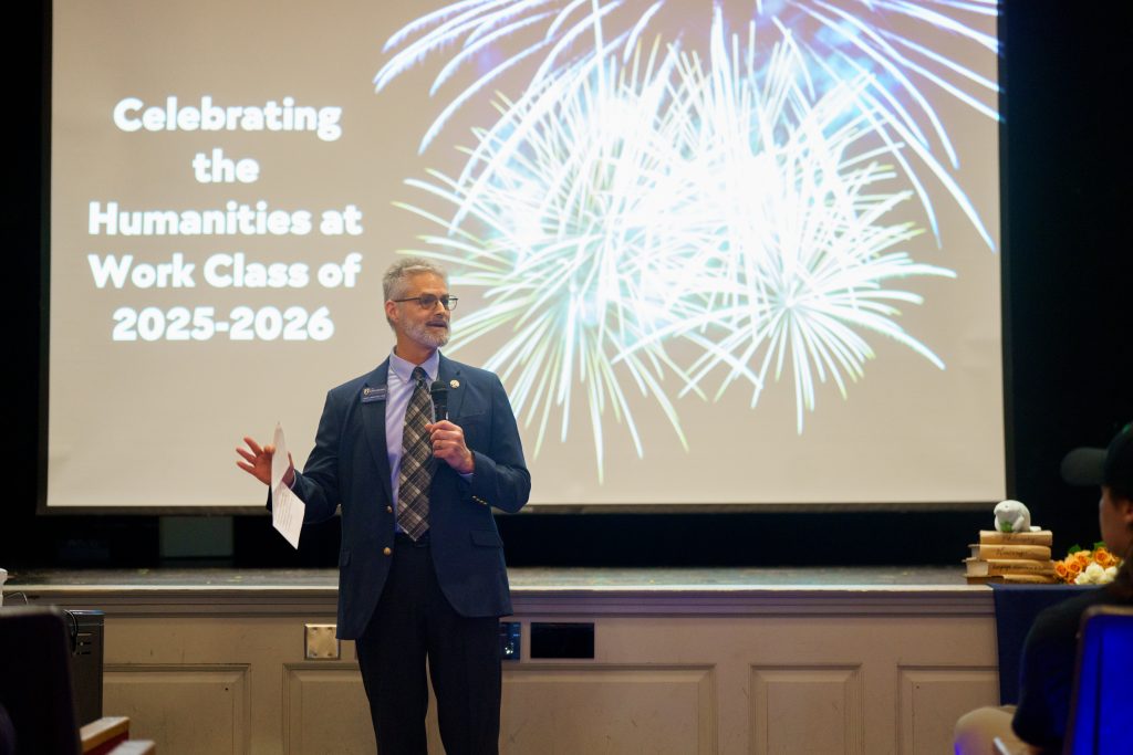 Dr. Alan Boyette stands in front of a projected screen with the words "Celebrating the Humanities at Work Class of 2025-26" with and an image of fireworks.