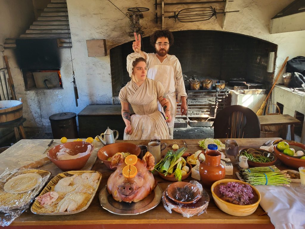 Two workers in replicas of colonial outfits strike fun poses, as they stand behind a table covered with recipes that would have been made in the 18th century. 