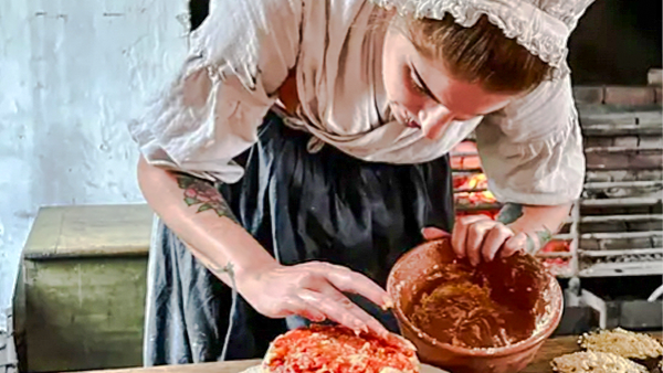A woman dressed in 18-th century period clothing prepares a dish in a historic kitchen.