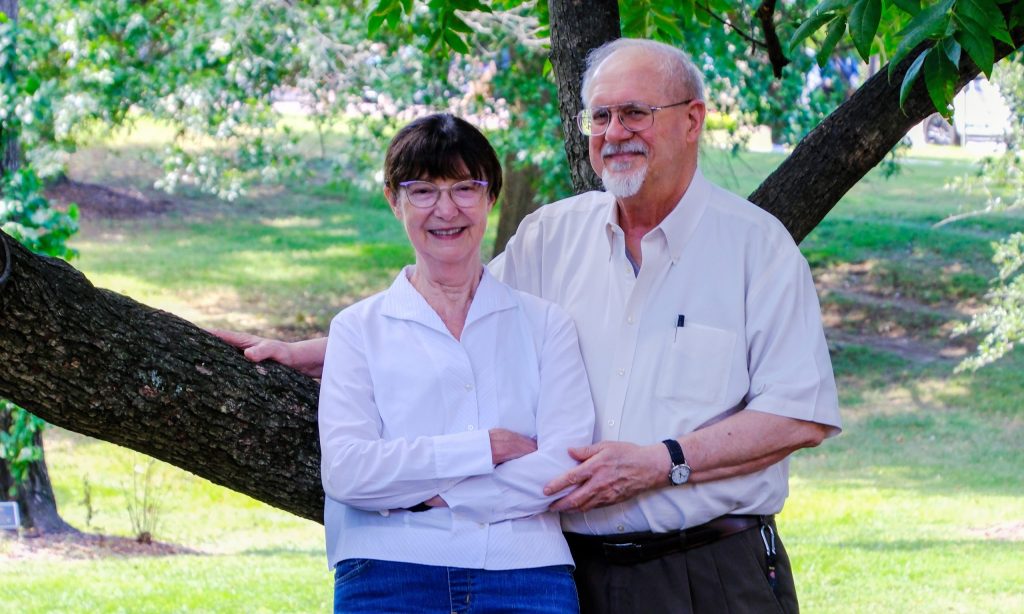 Laura and Christopher Tew hold hands beside a tree.